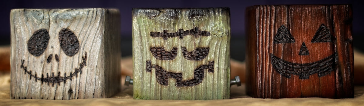 A close-up of three Halloween-themed, wooden tealight holders. The one on the left is white, with a Jack Skellington face. The one in the middle is light green, with a Frankenstein's Monster face. The one on the left is orange, with a pumpkin face. Shown on a purple and gold background.